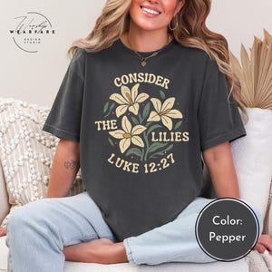 May include: Pepper-colored t-shirt featuring a floral design with the text "CONSIDER THE LILIES LUKE 12:27". The design includes cream-colored lilies and green leaves. The shirt is a relaxed fit.