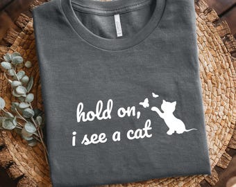 May include: A dark gray t-shirt with the text "hold on, i see a cat" in white script. A white cat silhouette reaches for butterflies. The shirt is folded on a woven surface.