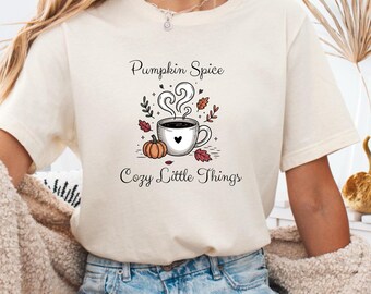 May include: A cream-coloured t-shirt with the words "Pumpkin Spice Cozy Little Things" and a graphic of a coffee cup with steam, a pumpkin, and autumn leaves. The design is in black, orange, and red.
