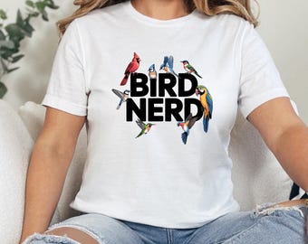 May include: White t-shirt with the text "BIRD NERD" in black, surrounded by colorful illustrations of various birds. The birds include a red cardinal, bluebirds, and a parrot.
