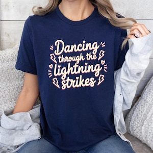 Pode incluir: Camiseta azul marinho com a frase "Dancing through the lightning strikes" em um design rosa e branco. O gráfico inclui raios e corações. A camiseta é de gola redonda e manga curta.