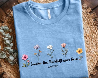 Könnte beinhalten: Hellblaues T-Shirt mit dem Text "Seht, wie die Lilien wachsen" und Lukas 12,27. Das Shirt zeigt Aquarellillustrationen von fünf verschiedenen Wildblumen in Rosa, Weiß, Orange und Gelb.