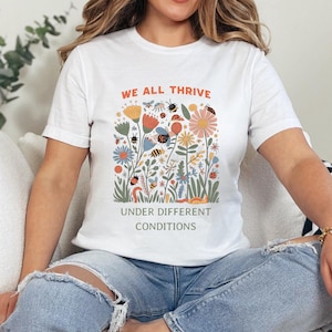 May include: White t-shirt with a colorful floral and insect graphic. The design includes flowers, bees, ladybugs, and the text "WE ALL THRIVE UNDER DIFFERENT CONDITIONS" in orange and gray.