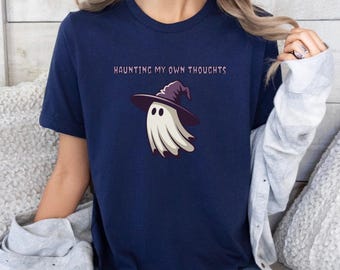 May include: Navy blue t-shirt with the phrase "HAUNTING MY OWN THOUGHTS" in purple text. The shirt features a cartoon ghost wearing a witch hat. The ghost is white with a purple hat and a brown brim.