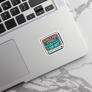 May include: A sticker with the text "VITALS ARE STABLE I AM NOT" in a retro-style design. The sticker features a teal and pink color scheme, with a monitor graphic and a heartbeat line.