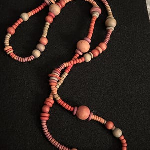 Op de afbeelding: Een lange ketting bestaande uit diverse houten kralen in tinten rood, oranje en beige. De kralen vari&euml;ren in grootte en vorm, waaronder kleine schijven en grotere bollen, geregen om een uniek accessoire te cre&euml;ren.