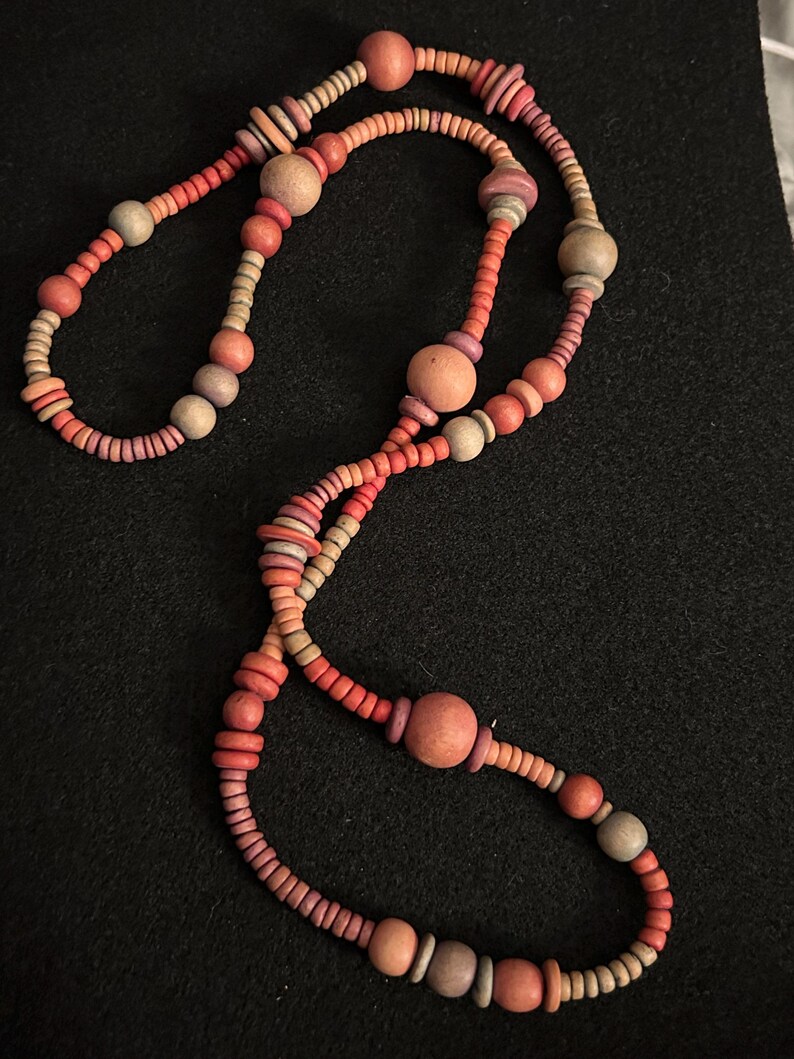Op de afbeelding: Een lange ketting bestaande uit diverse houten kralen in tinten rood, oranje en beige. De kralen vari&euml;ren in grootte en vorm, waaronder kleine schijven en grotere bollen, geregen om een uniek accessoire te cre&euml;ren.