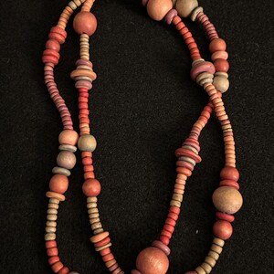 Op de afbeelding: Een meerstrengs ketting met houten kralen in rood, oranje en bruine tinten. De kralen vari&euml;ren in grootte en vorm, waaronder ronde en schijfvormige elementen. De ketting wordt gepresenteerd tegen een zwarte achtergrond.