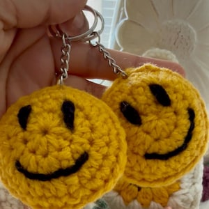 May include: Two yellow crocheted smiley face keychains with black eyes and smiles. The keychains are attached to silver metal key rings and chains. The background is a soft, blurred image of a white and pink floral design.