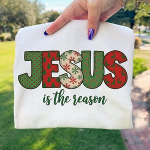 Puede incluir: Una camiseta blanca con la palabra JESUS en letras grandes y estampadas con motivos navideños rojos y verdes, incluyendo árboles, copos de nieve y bastones de caramelo. Debajo, el texto is the reason en cursiva verde oscuro.