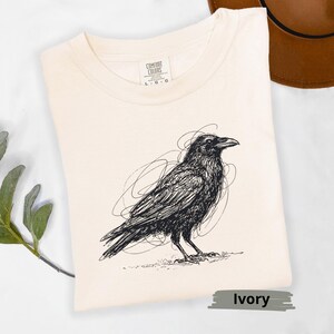 May include: Ivory-colored t-shirt featuring a detailed black raven illustration. The raven is depicted in a scribbled style, with a focus on its form and texture. The shirt is made of a soft material and is labeled "Comfort Colors".