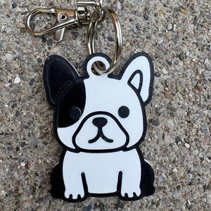Puede incluir: Un llavero de bulldog francés en blanco y negro con un mosquetón y un anillo plateados. El perro tiene una mancha negra sobre un ojo y orejas negras. El llavero está hecho de un material duradero.