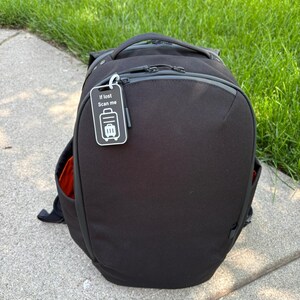 Puede incluir: Mochila negra con una etiqueta de equipaje que dice "If lost Scan me". La mochila tiene forma redondeada, un asa superior y múltiples compartimentos con cremallera. La etiqueta presenta un icono de maleta. La mochila está sobre una superficie de hormigón junto a hierba verde.