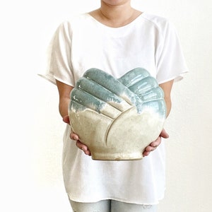 May include: A ceramic vase shaped like two hands clasped together. The vase is glazed in a light blue and white color scheme.