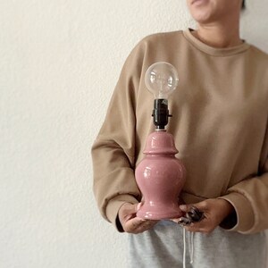 May include: A pink ceramic table lamp with a clear glass light bulb. The lamp is being held by a person wearing a brown sweater and gray shorts.