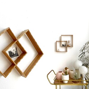 May include: Wooden wall shelves in a diamond and square design. The diamond shelf holds a black and white photograph. The square shelves display a small hourglass and figurines. A gold bar cart holds vases, plants, and globe lights.
