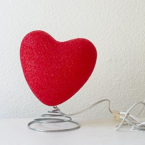 May include: A bright red heart-shaped lamp with a textured, granular surface. The heart is mounted on a silver-colored metal coil spring base. A white electrical cord with a plug extends from the base, trailing to the right side of the object.