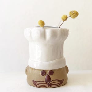 May include: A ceramic vase shaped like a chef's head, featuring a white hat and a brown face with a mustache. The vase holds a few yellow dried flowers. The design is whimsical and decorative.