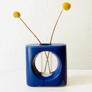 Puede incluir: Un jarrón de cerámica azul cobalto con un recorte circular, que contiene un tubo de vidrio. Se exhiben dos flores amarillas de billy button. El jarrón tiene un diseño geométrico moderno. El jarrón mide unos 15 cm de alto.