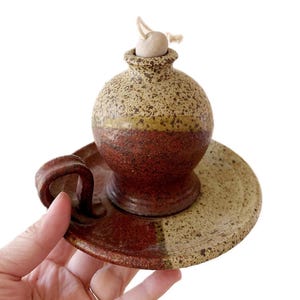 May include: A handmade ceramic oil lamp with a brown and beige speckled glaze. The lamp has a round body with a small handle and sits on a matching saucer.