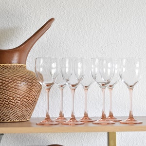 May include: A set of seven clear glass wine glasses with pink stems sit on a wooden shelf. A brown wicker pitcher with a woven design sits on the shelf behind the glasses.