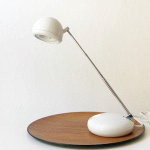 May include: A white desk lamp with a rounded head and a long, silver arm. The lamp sits on a wooden tray with a white base. The lamp is plugged in with a white cord.
