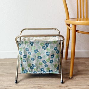May include: A vintage magazine rack with a chrome frame and a floral fabric liner. The fabric features a pattern of blue and green flowers on a white background. A wooden chair is next to the rack.