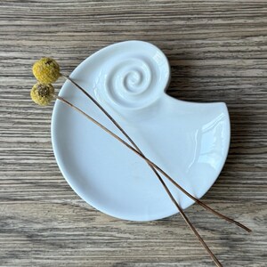 May include: A white ceramic seashell-shaped dish with a spiral design. Two dried yellow flower heads and two thin brown stems rest on the dish. The dish is on a wooden surface.