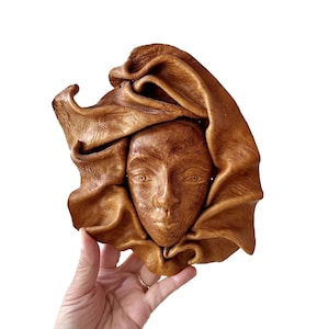 May include: A brown leather mask with a face sculpted into the leather. The mask is shaped like a head with a draped piece of leather over the top.