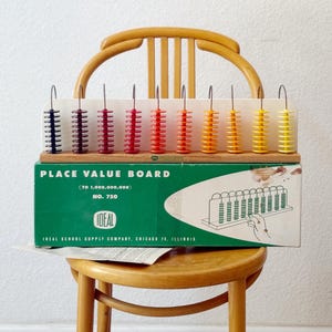 May include: A vintage Place Value Board with colorful plastic beads, displayed on a wooden chair. The board, labeled "IDEAL," has a green and white box with text. The beads are in shades of blue, red, orange, and yellow.