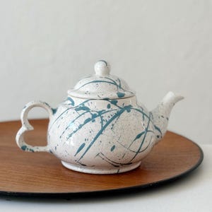 May include: A white ceramic teapot with a lid, adorned with teal paint splatter. The teapot features a heart-shaped handle and a spout. It sits on a round wooden tray.