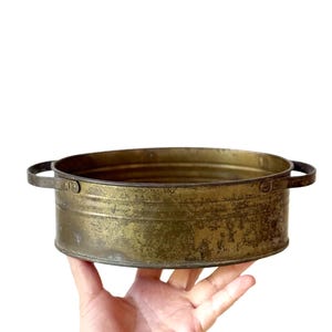 May include: A vintage, round metal container with two small handles. The container has a weathered, tarnished brass finish, showing signs of age and use. The container is held in a hand against a white background.