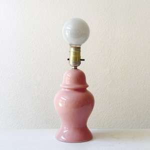 May include: A table lamp with a glossy, light pink ceramic base shaped like a ginger jar. The lamp features a metallic gold-toned socket and a single, round, frosted white light bulb positioned at the top, without a shade or additional cover.