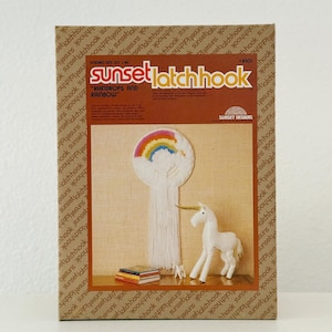 May include: A Sunset Latch Hook kit box featuring a finished project of a rainbow and cloud design with a unicorn figurine. The box has the text "RAINDROPS AND RAINBOW" and "FINISHED SIZE 10 x 14". The kit includes a small unicorn figurine.