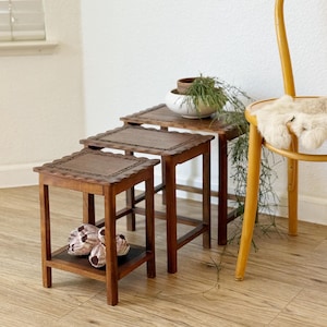 May include: A set of three wooden nesting tables with a dark brown finish. Each table has a square top with a decorative edge and a lower shelf. The tables are arranged in a nested configuration, with the smallest table inside the largest.
