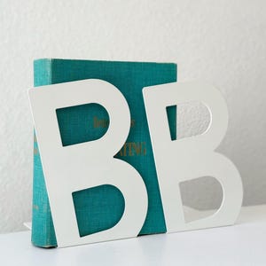 May include: White metal bookends in the shape of the letter "B" stand in front of a teal book. The bookends are on a white surface, with a white wall in the background. The bookends are a decorative item.