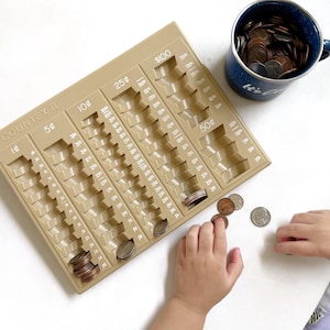 May include: A tan plastic coin sorter labeled "COUNTEX II" with slots for different coin denominations. A blue enamel mug filled with coins sits nearby. A child's hand is placing coins into the sorter.