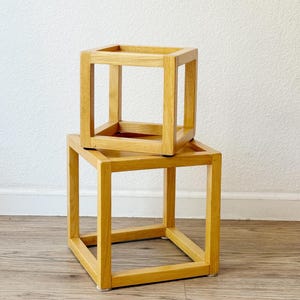 May include: Two stacked light brown wooden cube frames constructed from square beams. The smaller cube frame is positioned directly on top of the larger cube frame. Both frames feature an open design with four vertical posts and horizontal supports.