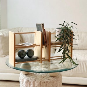 May include: A light wood bookshelf with two shelves, holding books and small items. Two dark green spheres sit on a rectangular marble base. The bookshelf is on a glass-topped table with a stone base, with a plant and a mirror in the background.