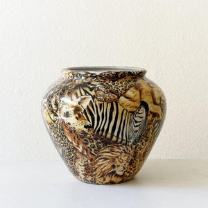 May include: A ceramic vase with a rounded body and a wide opening, featuring a detailed all-over print of various wild animals including a zebra, a lion, and leopard spots in shades of brown, black, tan, and white against a glossy finish.
