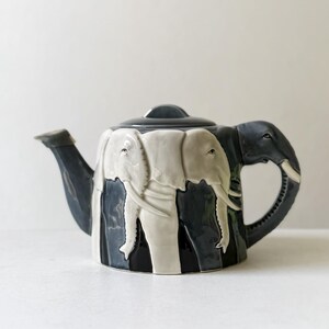 May include: A ceramic teapot featuring a repeating relief pattern of white and dark gray elephants around the body. The handle is shaped like an elephant head with tusks, and the spout is dark gray. It includes a matching dark gray lid.