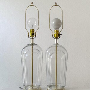 May include: Two clear glass table lamps with a classic design. Each lamp features a clear, rounded base, a gold-colored metal stem, and a gold-colored metal lamp holder. One lamp has a clear light bulb, and the other has a white light bulb.