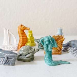 May include: A collection of small ceramic figurines featuring a turquoise mermaid, an orange seahorse, a green turtle, two grey whales, a white sailboat, a yellow castle, a light blue lighthouse, and an orange shell with the text MADE IN ENGLAND.