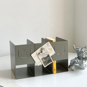 May include: A gray metal desk organizer with four compartments. Two vintage photographs are clipped to the front. A small yellow book and a gray decorative sculpture are also present.