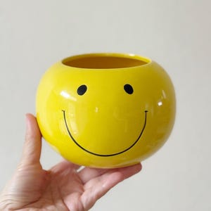 May include: A round, yellow ceramic smiley face planter. The planter has a wide opening and a black smiley face design. The planter is held in a hand against a neutral background.