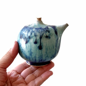 May include: A small, round, blue and green ceramic teapot with a cork stopper. The teapot has a unique, hand-painted design with blue and green swirls and drips.