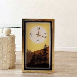 May include: A gold framed clock with a yellow and brown forest scene. The clock face has black Roman numerals and black hands.