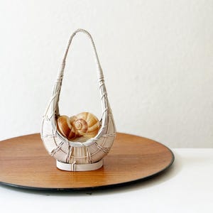 May include: A small, white woven basket with a curved handle, holding two large, brown and tan seashells. The basket sits on a round, wooden tray with a dark rim. The background is a plain white wall.