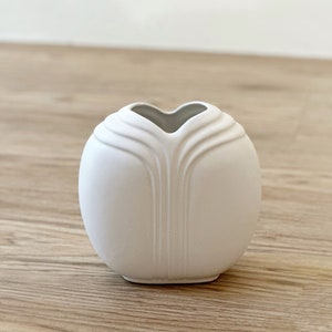 May include: A white ceramic vase with a heart-shaped opening and a smooth, rounded design. The vase has a subtle texture and a matte finish.