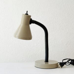 May include: A beige desk lamp with a black, flexible neck and a round base. The lamp has a conical shade and a power cord with a plug. The lamp is designed for desk use.